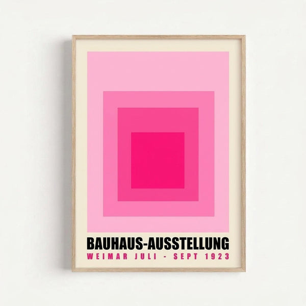 May include: Framed poster with concentric pink squares in varying shades from light to deep magenta, creating a geometric gradient effect. Black text below reads "BAUHAUS-AUSSTELLUNG" with pink text "WEIMAR JULI - SEPT 1923". Light wood frame with white matting, mounted on white wall with subtle shadow.