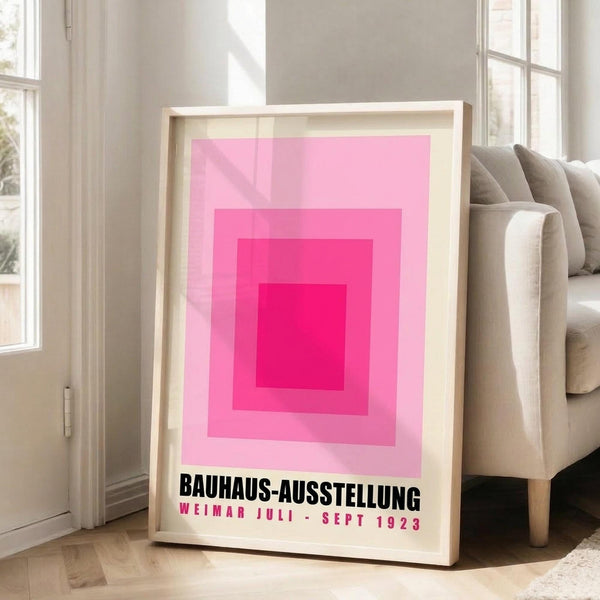 May include: Framed art print with concentric pink and magenta squares creating gradient effect from deep pink center to pale pink edges on cream background. Black text reads "BAUHAUS-AUSSTELLUNG WEIMAR JULI - SEPT 1923" below geometric design. Print in light wood frame leans against white wall next to beige upholstered sofa in bright, minimalist living room with large windows and natural light.