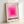 May include: Framed poster featuring concentric pink squares in gradient shades from deep magenta center to pale pink, with black text "BAUHAUS-AUSSTELLUNG" and pink text "WEIMAR JULI - SEPT 1923" below. Light wood frame with white matting, leaning against white wall on white surface.