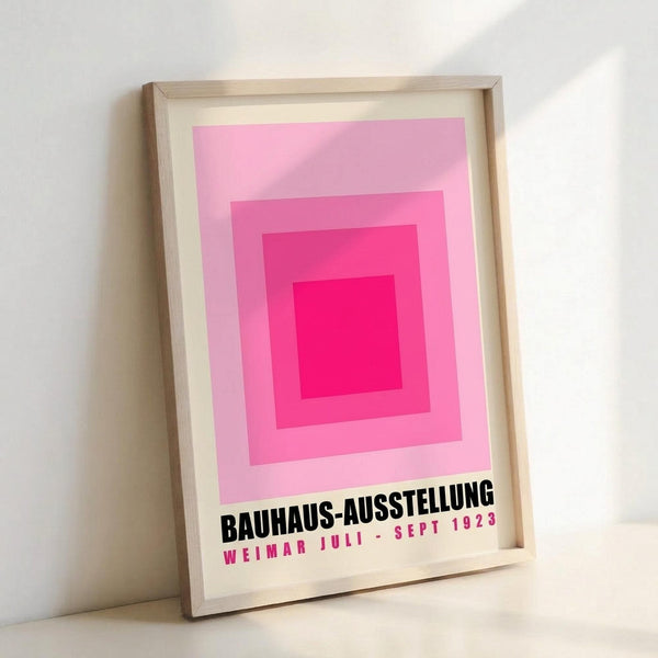May include: Framed poster featuring concentric pink squares in gradient shades from deep magenta center to pale pink, with black text "BAUHAUS-AUSSTELLUNG" and pink text "WEIMAR JULI - SEPT 1923" below. Light wood frame with white matting, leaning against white wall on white surface.