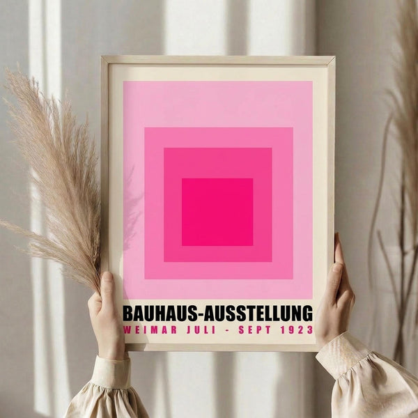 May include: Framed poster with pink gradient squares in concentric pattern, from light pink outer border to bright magenta center. Black text below reads "BAUHAUS-AUSSTELLUNG" and "WEIMAR JULI - SEPT 1923" in red letters. Person in beige sweater holds wooden frame alongside dried pampas grass against neutral curtained background.