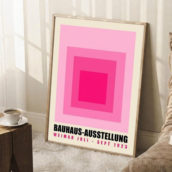 May include: A framed print featuring concentric pink squares in gradient shades from light to bright magenta, with black German text below reading "BAUHAUS-AUSSTELLUNG WEIMAR JULI - SEPT 1923". The wooden frame leans against white wainscoting beside a small wooden side table with white coffee cup and saucer, on a textured cream rug next to a beige knit throw blanket.