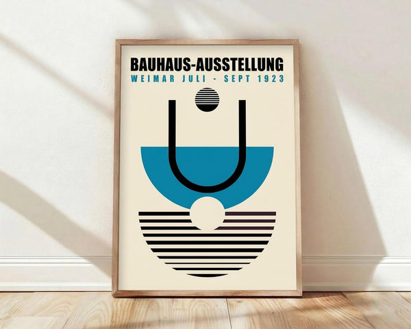 May include: A framed vintage-style poster with cream background featuring geometric Bauhaus design elements in black and teal blue. Bold black text reads "BAUHAUS-AUSSTELLUNG WEIMAR JULI-SEPT 1923" above abstract shapes including a striped circle, curved lines, and semicircular forms. The wooden frame leans against a white wall on light wooden flooring with natural window light creating soft shadows.