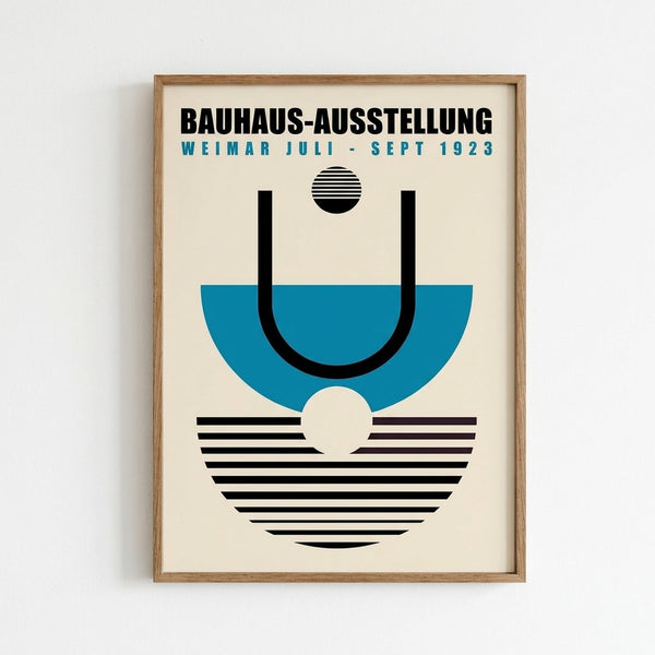 A framed poster with geometric design featuring black text "BAUHAUS-AUSSTELLUNG WEIMAR JULI - SEPT 1923" at top, a blue semi-circle with black curved lines forming a "U" shape, small black striped circle above, and black and white striped semi-circle below with cream circle accent. Clean modernist aesthetic in light wood frame against white wall.