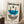 May include: Framed Bauhaus exhibition poster leaning against white wall corner on textured cream rug. Features geometric design with blue semicircle, black curved lines, striped circles in black and white. Text reads "Bauhaus-Ausstellung Weimar Juli-Sept 1923" in black and teal. Light wood frame, coffee cup on wooden block nearby, beige throw blanket visible. Clean modernist aesthetic with natural lighting casting soft shadows.