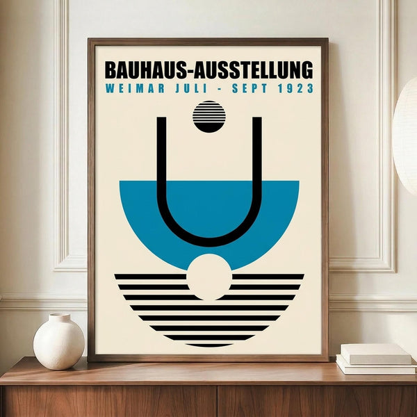 May include: Framed poster with geometric Bauhaus design featuring blue semicircle, black curved lines, striped circles and semicircle elements on cream background. Text reads "Bauhaus-Ausstellung Weimar Juli-Sept 1923" in black and blue letters. Wooden frame sits on mid-century credenza next to white ceramic vase and books in minimalist room with white walls and molding.