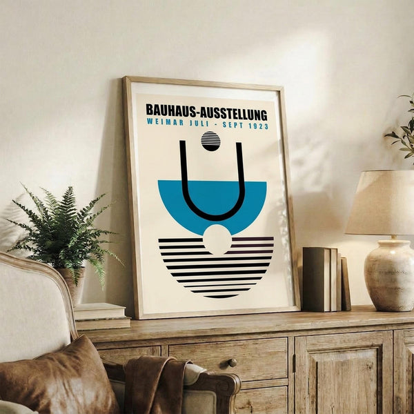 May include: Framed Bauhaus exhibition poster with geometric design in blue, black and white featuring abstract shapes and striped patterns, displayed on rustic wooden console table alongside fern plant, books, and ceramic pottery in cozy living room setting with beige armchair
