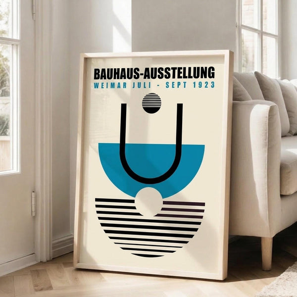 May include: Framed poster with cream background featuring geometric design in blue and black. Abstract composition shows curved and striped elements with small circles, displaying Bauhaus-style minimalist artwork. Light wood frame leans against white wall near beige upholstered chair in bright room with large windows.