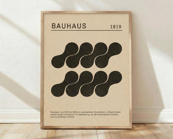 May include: Framed Bauhaus-inspired poster with cream background featuring black organic blob shapes arranged in two rows of four. The design includes "BAUHAUS 1919" text at top and German descriptive text at bottom. Light wood frame sits on wooden floor against white wall with natural lighting and shadows.