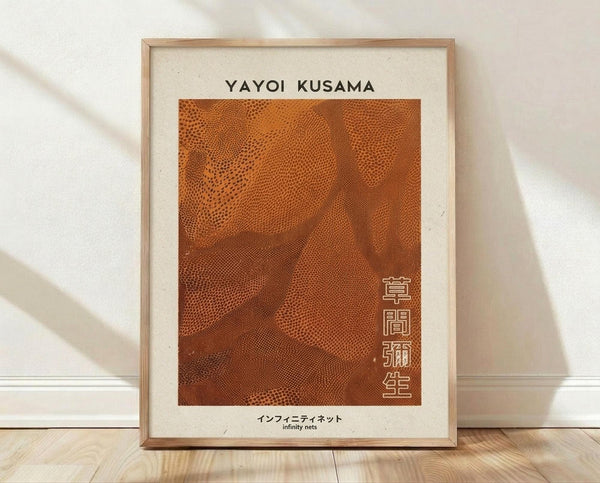 May include: Framed art print leaning against white wall on wooden floor. Light wooden frame contains cream-colored poster with "YAYOI KUSAMA" text at top, orange-brown dotted pattern artwork resembling flowing nets or waves, and Japanese characters vertically aligned on right side. Bottom shows "infinity nets" text. Natural lighting creates soft shadows on textured white wall background.
