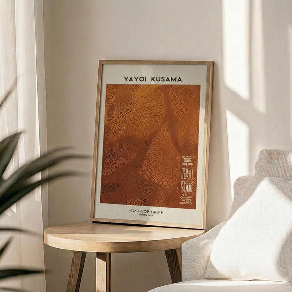 May include: Framed art print with orange dotted pattern and Japanese text displayed on wooden side table next to beige upholstered chair. Natural lighting creates soft shadows across neutral-toned room with plant leaves visible in corner. Print features warm terracotta background with small repeated dots creating flowing organic shapes, presented in thin light wood frame.