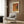 May include: Cream-colored poster with orange and pink abstract design featuring Japanese text, displayed in light wood frame on wood paneled wall. White upholstered armchair with geometric patterned pillow sits below artwork, alongside brass floor lamp and dark round side table with open book.