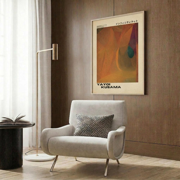 May include: Cream-colored poster with orange and pink abstract design featuring Japanese text, displayed in light wood frame on wood paneled wall. White upholstered armchair with geometric patterned pillow sits below artwork, alongside brass floor lamp and dark round side table with open book.