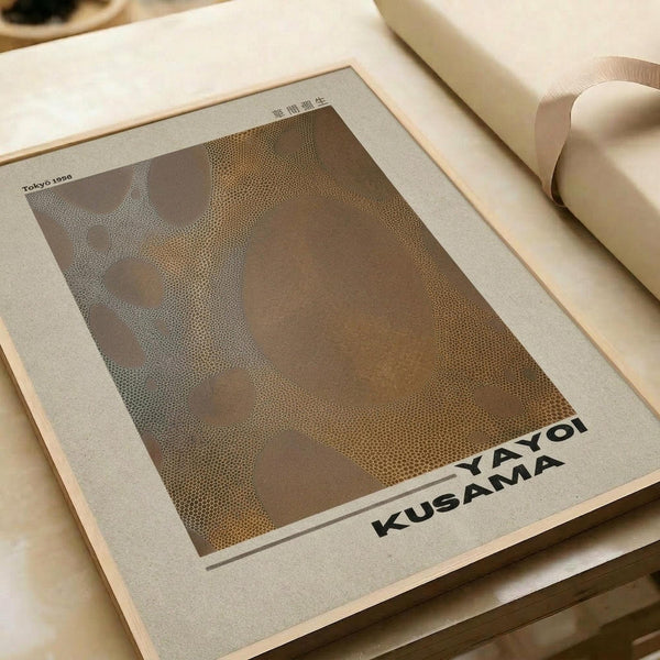 Minimalist art print featuring abstract organic shapes in brown and beige tones created with dotted stippling technique. The poster displays "YAYOI KUSAMA" text at bottom and "Tokyo 1998" in upper left corner with Japanese characters. Print has cream colored border and is photographed on neutral surface next to rolled paper tube, showing gallery-style presentation.