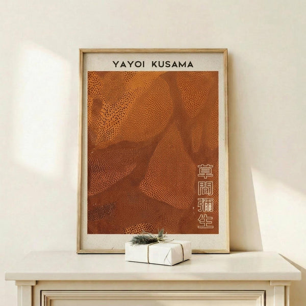 May include: Framed poster with orange dotted pattern artwork in light wood frame, leaning against white wall on light wooden surface next to small wrapped gift with evergreen sprig. The poster features flowing organic shapes created with tiny dots in warm orange and brown tones, with vertical Japanese characters in bottom right corner.