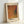 May include: Framed art print leaning against white wall showing abstract composition with warm orange, brown and purple geometric shapes plus small teal circle on textured dotted background. Japanese text appears at top, artist name at bottom in black letters on cream border. Light wood frame casts soft shadow.