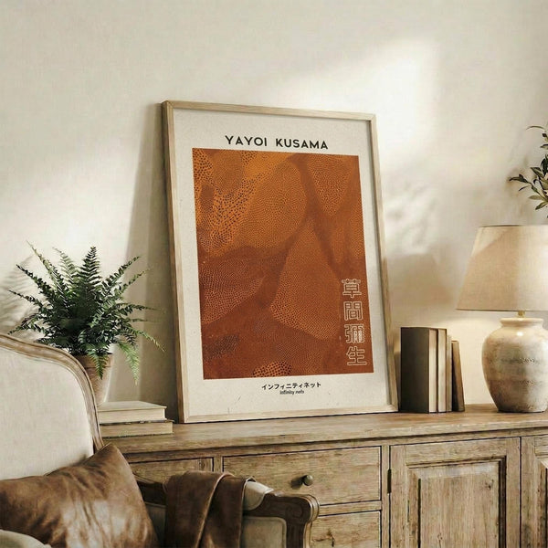 May include: Orange abstract art print with dotted pattern displayed in light wood frame on rustic wooden sideboard. Room setting shows beige armchair, fern plant, stacked books, ceramic vase, and soft natural lighting against white wall.