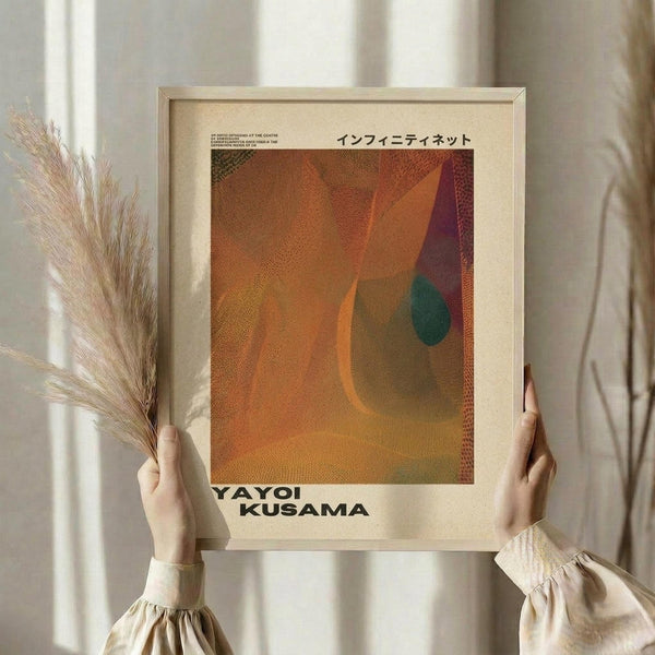 May include: Hands holding a framed art print featuring abstract organic shapes in warm orange, rust, and burgundy tones with a teal accent, displayed against white curtains. Person wears a cream-colored blouse with gathered sleeves and holds dried pampas grass in one hand. The artwork has a textured, dotted pattern throughout and Japanese text at the top.