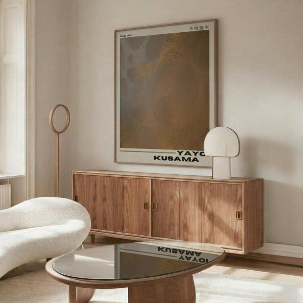 May include: Modern living room with walnut credenza displaying framed abstract art poster in earthy brown and olive tones with dotted pattern. White curved chair, round glass coffee table, and floor lamp create mid-century aesthetic. Poster shows geometric stippled design in warm neutral colors within light wood frame.