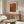 May include: Orange textured art print with polka dot pattern and Japanese text hanging above wooden sideboard in modern living room. White curved chair and glass coffee table with colorful abstract design visible. Warm wood tones and neutral walls create minimalist Scandinavian-style interior setting.