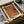 May include: Abstract art print with warm orange, brown and purple gradients and a small teal circle, displayed in black frame on cream mat. Print shows textured dotted pattern throughout. Packaged in brown cardboard box with white tissue paper on white surface alongside wooden dowels.