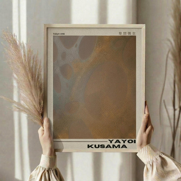 May include: Person in cream ruffle-sleeved top holding framed abstract art print featuring organic brown and tan dotted patterns. Light wood frame with white matting, Japanese text visible at top. Dried pampas grass stems held in other hand against soft neutral curtained background with natural window lighting.