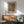 May include: Modern living room with Yayoi Kusama exhibition poster in wooden frame hanging on white wall above mid-century wooden credenza. Poster features abstract brown and yellow dotted pattern with artist's name. Scene shows sage green upholstered armchair with wooden arms, white ceramic table lamp, and sunlight casting shadows across the minimalist space.