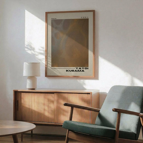May include: Modern living room with Yayoi Kusama exhibition poster in wooden frame hanging on white wall above mid-century wooden credenza. Poster features abstract brown and yellow dotted pattern with artist's name. Scene shows sage green upholstered armchair with wooden arms, white ceramic table lamp, and sunlight casting shadows across the minimalist space.