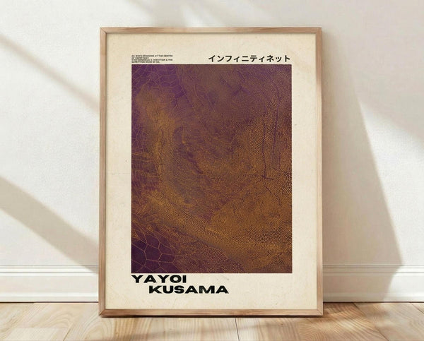 May include: Framed art print leaning against white wall on wooden floor. Light wood frame contains cream-colored poster with purple and brown textured pattern resembling organic cellular or net-like forms. Japanese text at top, "YAYOI KUSAMA" in bold black letters at bottom. Natural window light creates soft shadows on wall. Modern minimalist interior setting.