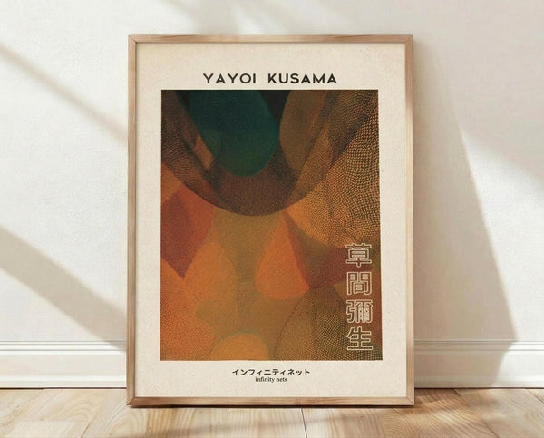 May include: Framed art print leaning against white textured wall on light wood floor. Artwork features abstract organic shapes in warm earth tones - orange, brown, rust, and dark green - with dotted texture patterns throughout. Natural wood frame surrounds cream matting. Text reads "YAYOI KUSAMA" at top, Japanese characters on right side, and "infinity nets" at bottom. Soft natural lighting creates diagonal shadows across the scene.