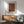 May include: Framed art print with purple and golden brown dotted pattern hanging above wooden sideboard. Mid-century modern interior with sage green armchair, white ceramic table lamp, and natural sunlight casting shadows on white wall. Warm wood tones throughout furniture pieces.