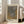 May include: Framed art poster leaning against white wall next to beige upholstered sofa in bright living room with large windows. The poster features abstract dotted patterns in grey and golden brown tones with Japanese text at top and artist name at bottom. Light wooden frame, approximately 2-3 feet tall, casting soft shadows on hardwood floor.