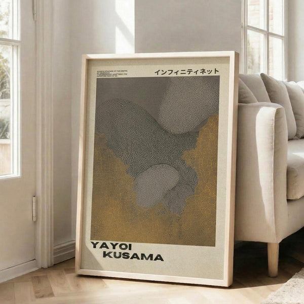 May include: Framed art poster leaning against white wall next to beige upholstered sofa in bright living room with large windows. The poster features abstract dotted patterns in grey and golden brown tones with Japanese text at top and artist name at bottom. Light wooden frame, approximately 2-3 feet tall, casting soft shadows on hardwood floor.