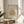 May include: Framed art print displaying golden botanical foliage design on soft pink and cream background in thin wooden frame, positioned on round wooden side table next to beige upholstered chair, with plant leaves visible in corner and natural window light creating shadows on neutral wall