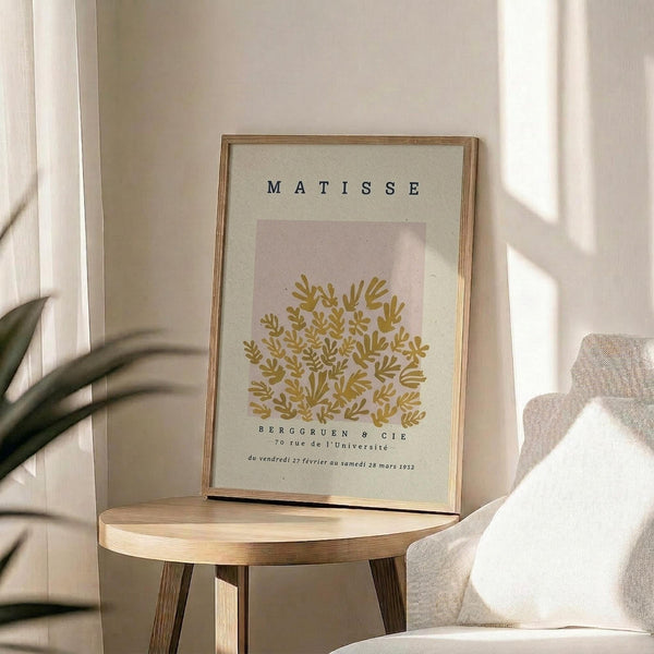 May include: Framed art print displaying golden botanical foliage design on soft pink and cream background in thin wooden frame, positioned on round wooden side table next to beige upholstered chair, with plant leaves visible in corner and natural window light creating shadows on neutral wall