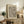 May include: Black and white abstract wavy line pattern poster with "Yayoi Kusama" text at top, displayed in light wood frame on rustic wooden sideboard. Room scene shows neutral tones with beige armchair, small potted fern, ceramic vase, and stacked books. Wall art features flowing curved parallel lines creating organic wave-like movement against cream background.