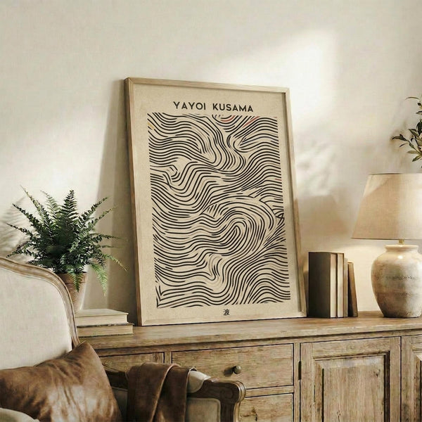 May include: Black and white abstract wavy line pattern poster with "Yayoi Kusama" text at top, displayed in light wood frame on rustic wooden sideboard. Room scene shows neutral tones with beige armchair, small potted fern, ceramic vase, and stacked books. Wall art features flowing curved parallel lines creating organic wave-like movement against cream background.