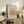 May include: Framed poster with cream background featuring golden botanical leaves on pink rectangle, displayed on rustic wooden sideboard alongside books, ceramic vases, and fern plant in neutral-toned living room setting