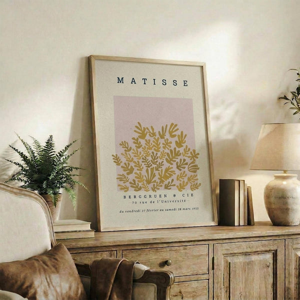 May include: Framed poster with cream background featuring golden botanical leaves on pink rectangle, displayed on rustic wooden sideboard alongside books, ceramic vases, and fern plant in neutral-toned living room setting