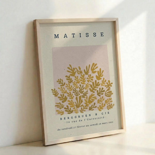 Framed vintage-style art print featuring golden botanical leaf motifs on a soft pink and cream background. The poster displays "MATISSE" at the top and "BERGGRUEN & CIE" with French exhibition text at the bottom. Set in a light wood frame against a white wall, the print has a minimalist, mid-century modern aesthetic with organic, hand-drawn plant forms clustered in the center.