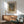 May include: Framed abstract art poster with organic dotted patterns in green, yellow and gray tones displayed above a wooden sideboard. Mid-century modern living room setting features a sage green armchair with wooden frame, white ceramic table lamp, and dramatic natural light casting shadows across white walls.