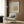 May include: Beige armchair with geometric pillow in modern living room with wood-paneled walls. Framed art poster shows golden botanical leaf design on pink background with "Matisse" text. Gold floor lamp and dark round side table with open book complete the neutral-toned interior scene.