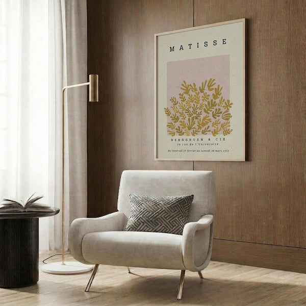 May include: Beige armchair with geometric pillow in modern living room with wood-paneled walls. Framed art poster shows golden botanical leaf design on pink background with "Matisse" text. Gold floor lamp and dark round side table with open book complete the neutral-toned interior scene.