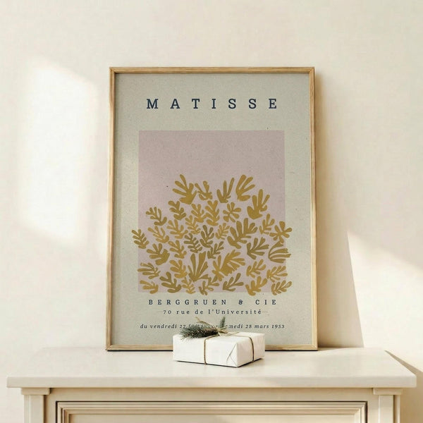 May include: Framed art print leaning against white wall on light wood surface. Features pink and cream background with golden-yellow botanical coral or seaweed-like design. Black text at top reads "MATISSE" with exhibition details below including "BERGGRUEN & CIE" and French venue information from 1953. Natural wood frame surrounds the poster. Small white wrapped gift with greenery sits in front. Soft natural lighting creates gentle shadows.