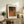 May include: Framed Yayoi Kusama-inspired abstract art print with warm orange, brown, and green organic shapes and dotted textures, displayed on rustic wooden sideboard alongside potted fern, ceramic vases, and books in cozy living room with beige armchair and neutral walls