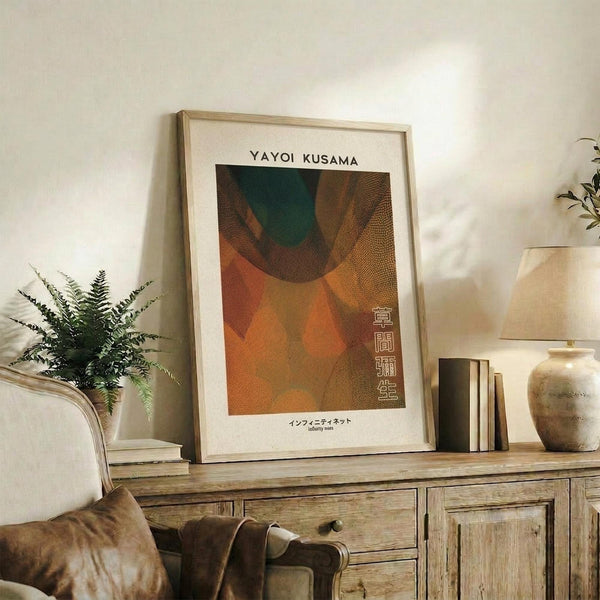 May include: Framed Yayoi Kusama-inspired abstract art print with warm orange, brown, and green organic shapes and dotted textures, displayed on rustic wooden sideboard alongside potted fern, ceramic vases, and books in cozy living room with beige armchair and neutral walls