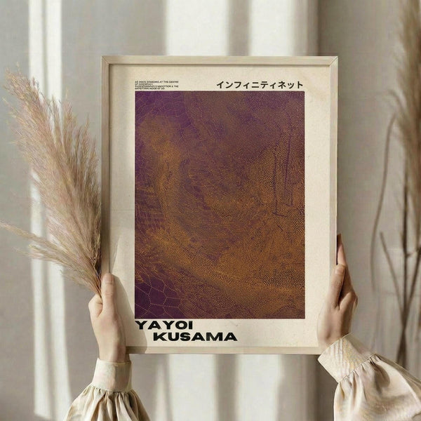 May include: Hands holding a framed art print with purple and orange dotted pattern resembling Kusama's Infinity Nets style, displayed against neutral curtains with dried wheat stalks nearby. The poster features Japanese text at top and "YAYOI KUSAMA" at bottom in bold letters on cream background within light wood frame.
