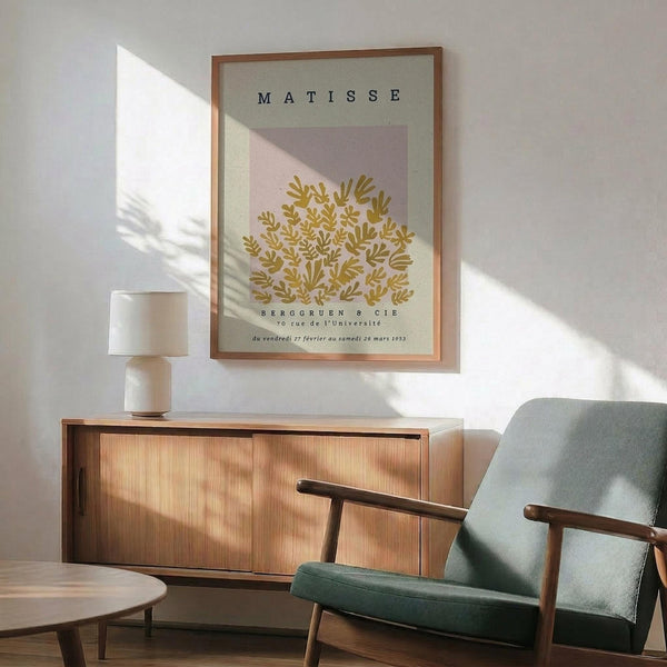 May include: Framed Matisse exhibition poster with golden botanical cutout design on pale background, displayed above wooden sideboard with ceramic lamp. Mid-century modern living room setting features sage green upholstered armchair with wooden frame, round coffee table, and natural sunlight casting shadows across white walls.