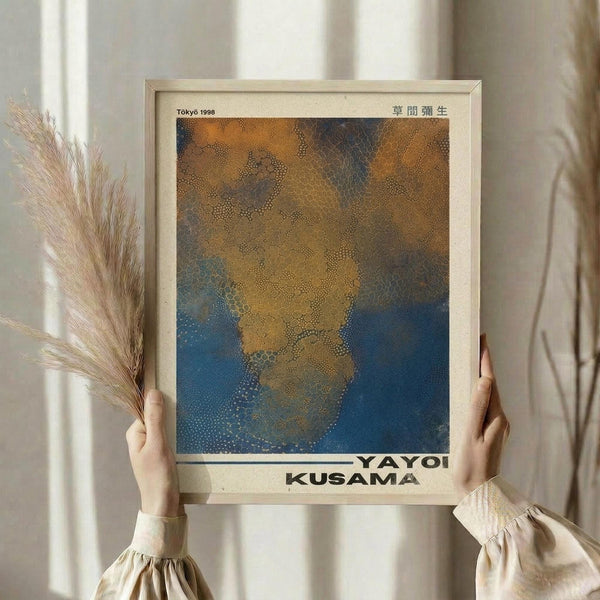 May include: Person holding framed art print featuring abstract dotted pattern in golden yellow, orange, and deep blue tones. Light wooden frame against neutral wall with dried wheat stalks visible. Person wears cream-colored blouse with gathered sleeves. Soft natural lighting creates warm, minimalist aesthetic.