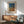 May include: Framed poster with abstract dotted pattern in blue, orange and brown tones displayed above wooden sideboard in modern living room. Green upholstered mid-century chair with wooden arms sits nearby, white ceramic table lamp on credenza, sunlight streaming across white walls creating geometric shadows.